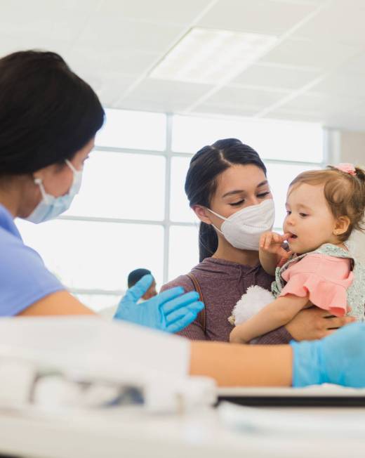 A female healthcare professional gestures as she talks to a young mother and the woman's baby. The nurse and mother are wearing protective face masks during the coronavirus outbreak.